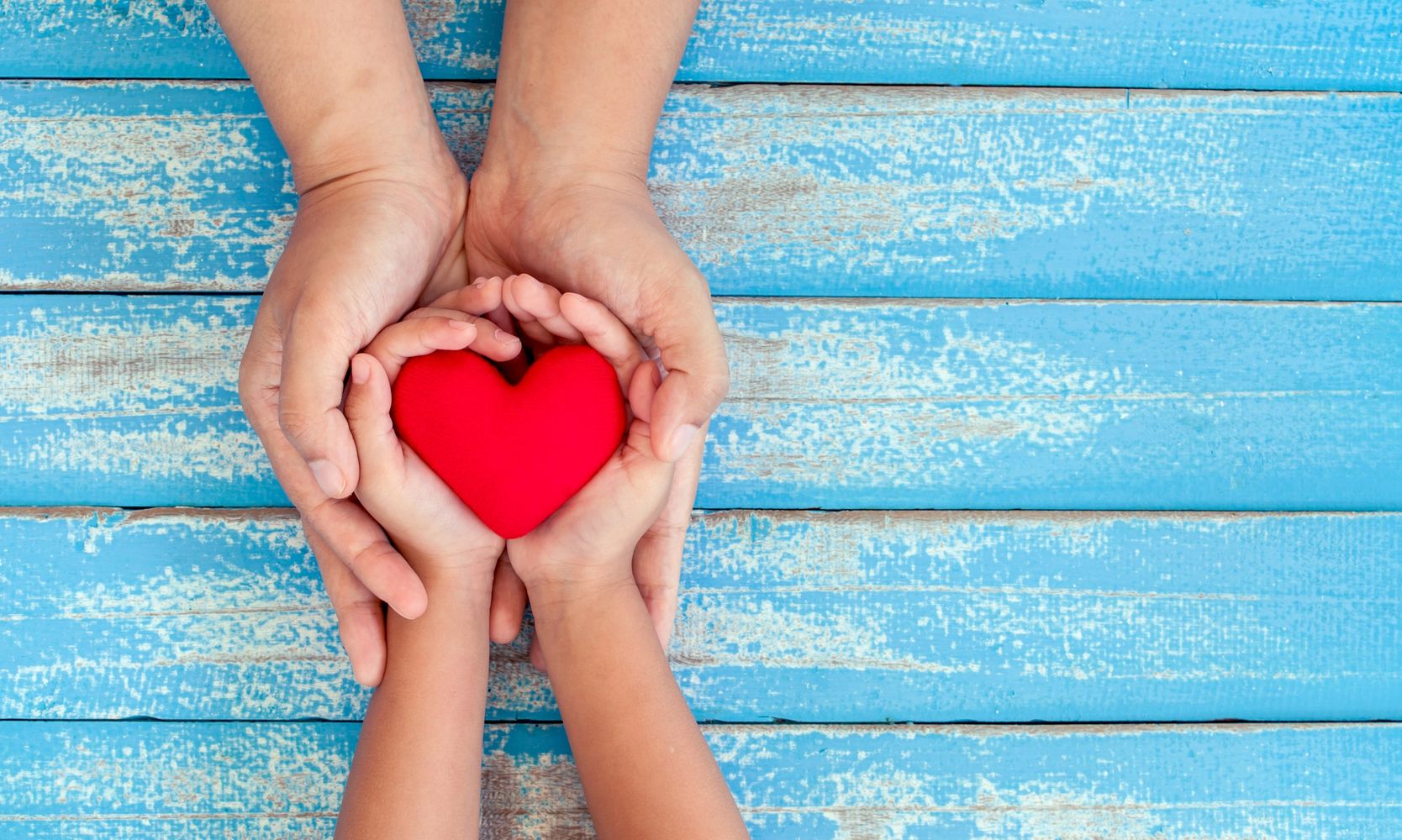 Adult and child hands holding a red heart against a blue wooden background.
