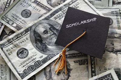 Graduation cap labeled 'SCHOLARSHIP' on a pile of US dollar bills.