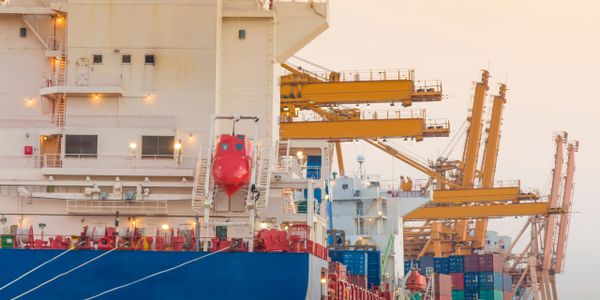 Cargo ship docked at a busy container port with cranes in the background.