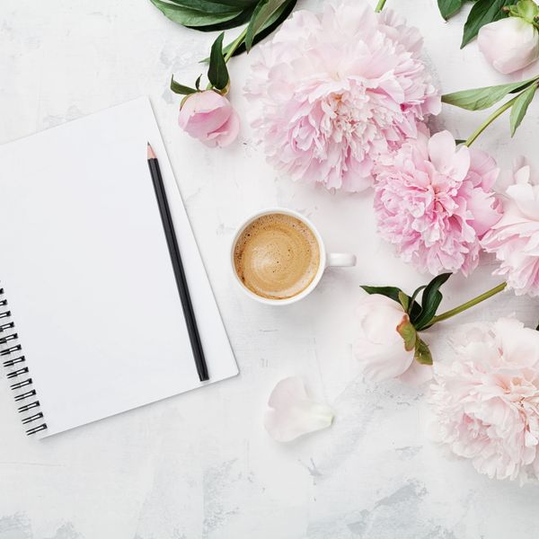 Notebook, pencil, coffee, and pink peonies on a white textured surface.