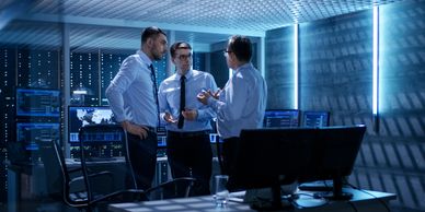 Three men in business attire discussing in a high-tech office with multiple monitors.