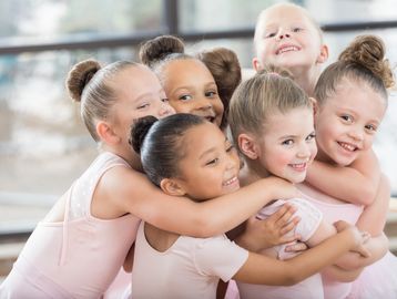 A group of six young girls in ballet attire hugging and smiling.