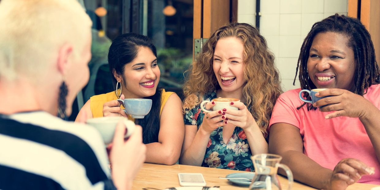 Midlife women are gathered around a table, holding cups of coffee, smiling, laughing, and talking.