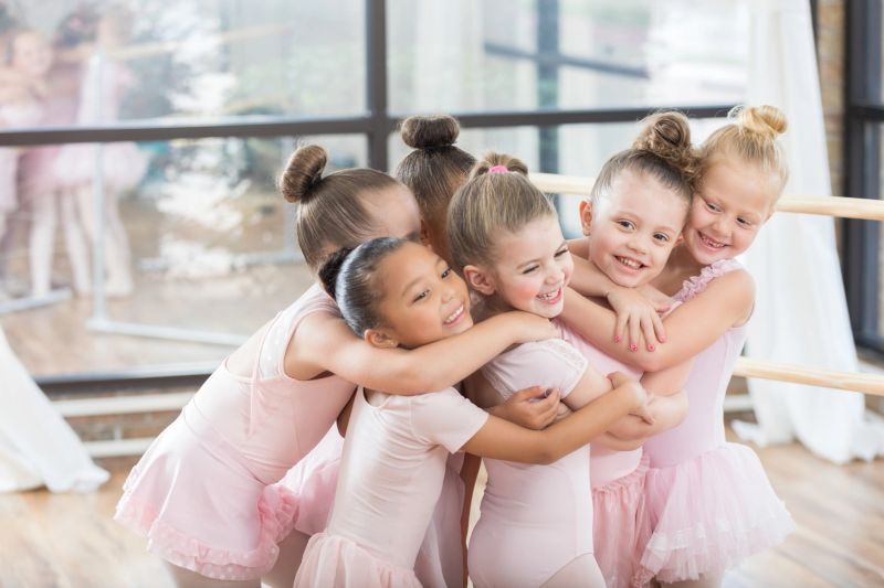 Happy group of diverse young ballerinas hug and are playful with one another after their ballet class. They are standing in front of a large window.