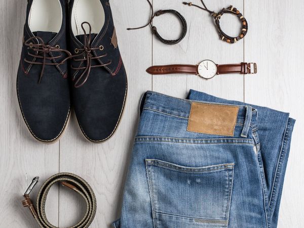 Casual men's outfit with shoes, jeans, belt, watch, and bracelets on a wooden floor.