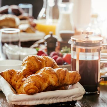 Fresh croissants in a basket with coffee and breakfast items in the background.
