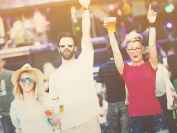 Three friends enjoying a lively outdoor music festival with drinks in hand.