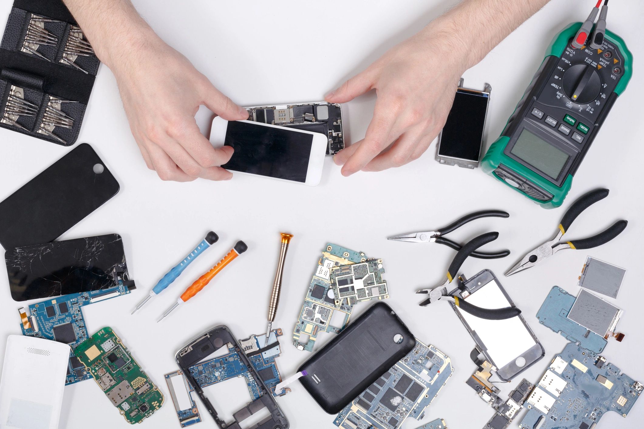 Hands repairing a disassembled smartphone with various tools and parts on a white surface.