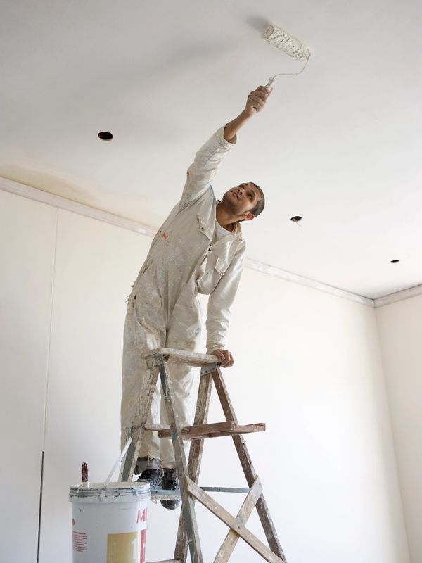 Painter on ladder rolling white paint on ceiling.