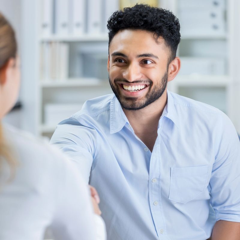 This is a closeup of an attractive young man smiling broadly as he shakes hands with his unrecognizable new female manager.  She has just offered him a job on the spot.