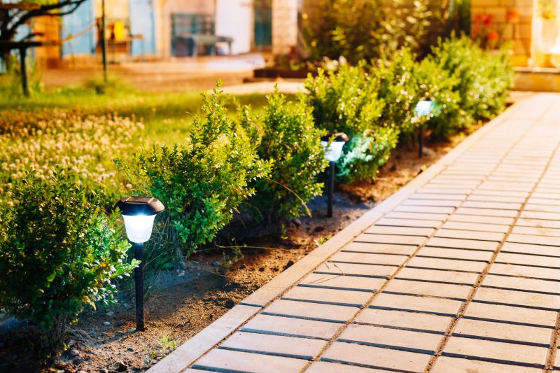 Night View Of Flowerbed With Violas Illuminated By Energy-Saving Solar Powered Lanterns Along The Path Causeway On Courtyard Going To The HouseBeautiful Small Solar Garden Light, Lanterns In Flower Bed. Garden Design. Solar Powered Lamp