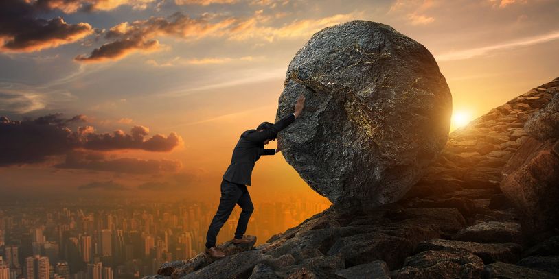 A man in a suit pushes a huge boulder uphill at sunset.