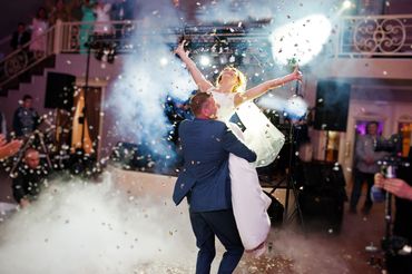 Groom lifts bride joyfully amidst confetti and fog at wedding celebration.