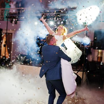 A groom lifts his joyful bride amidst confetti and smoke effects at their wedding celebration.