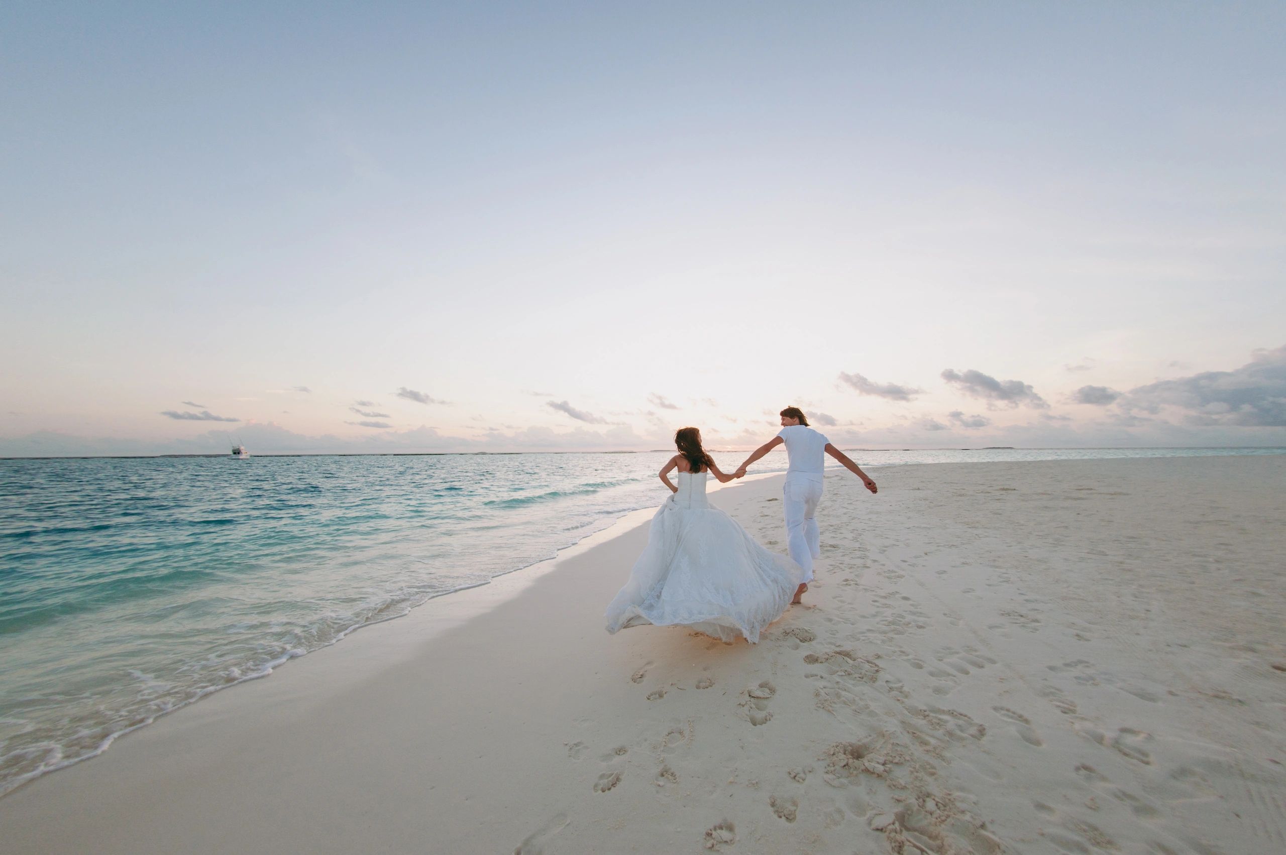 Couple in wedding attire walking hand in hand on a serene beach at sunset.