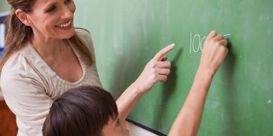 Teacher guiding a young student writing on a chalkboard.