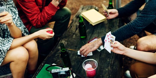 Friends playing cards around a rustic wooden table outdoors with drinks and books.