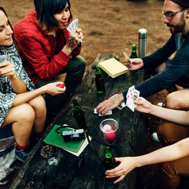 Friends playing cards outdoors around a wooden table with drinks.