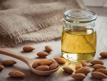 almonds on a table and in a wooden spoon with a clear glass filled with almond oil