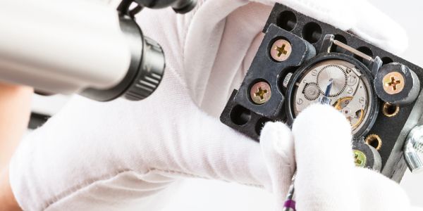 A gloved technician repairing a watch mechanism under a microscope.