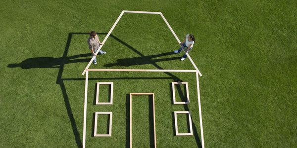 Two people holding wooden frames forming a house outline on grass.