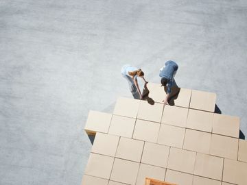 Two people assembling large cardboard boxes on a concrete surface.