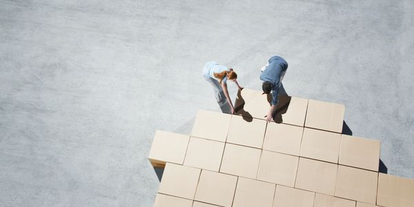 Two people arranging cardboard boxes together on a concrete floor.
