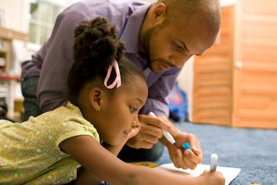 A father helps his daughter with drawing on paper on the floor.