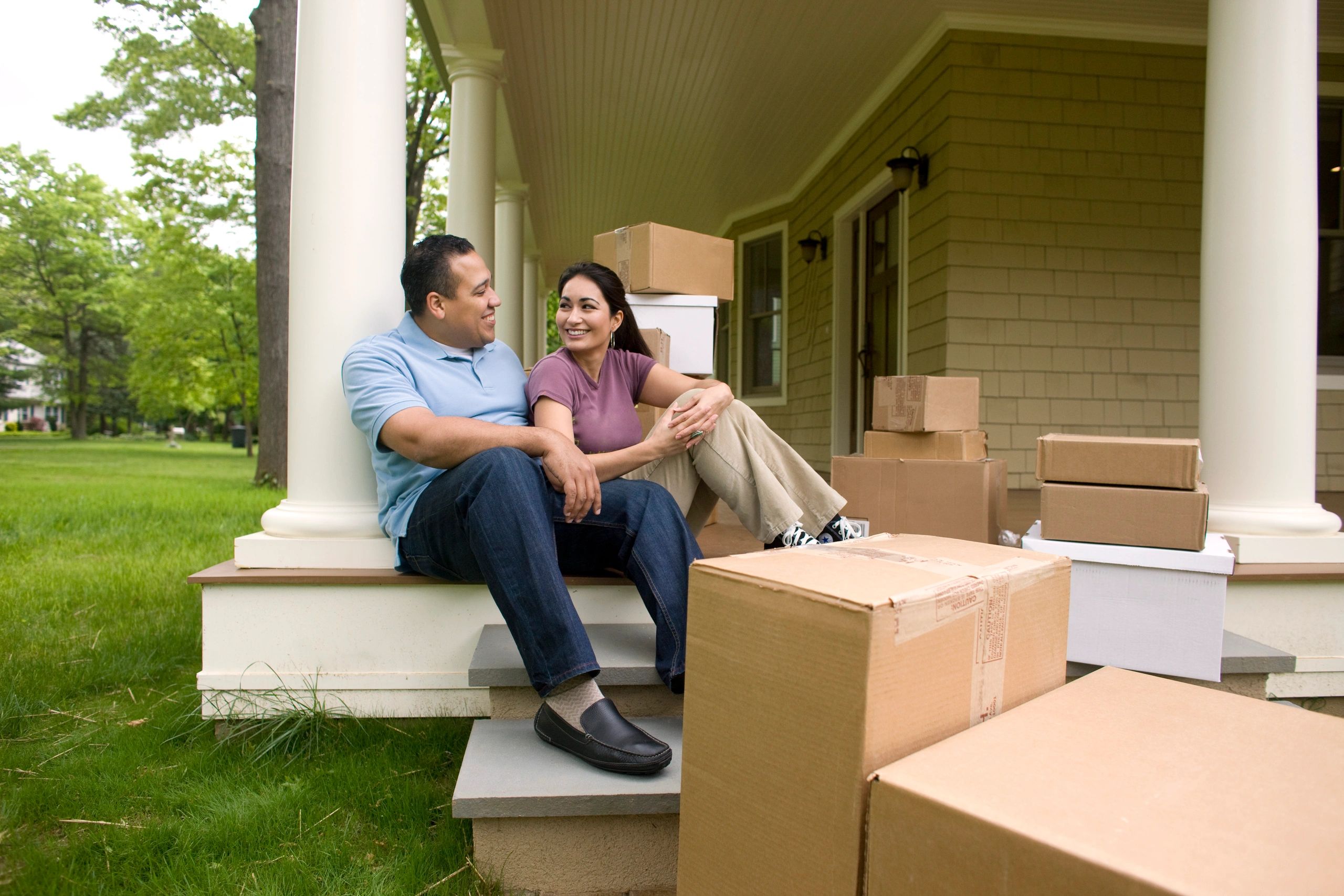 A smiling young couple sitting together on the front porch steps of a house, surrounded by cardboard