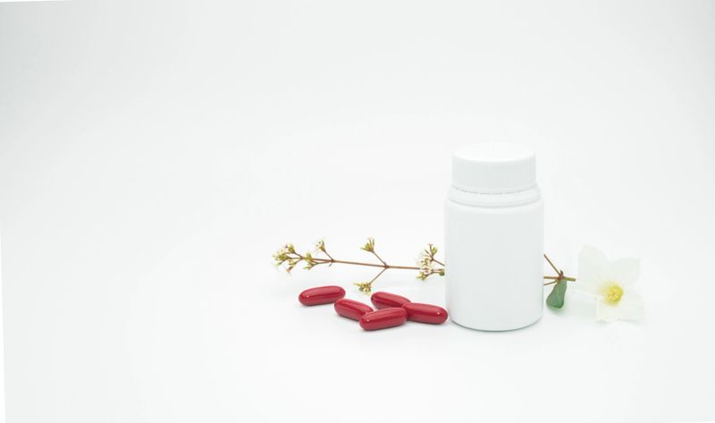 Red vitamin and supplement capsule pills with flower and branch with blank label plastic bottle on white background with copy space, just add your own text
