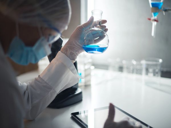 Scientist examining blue liquid in flask while using tablet in lab.