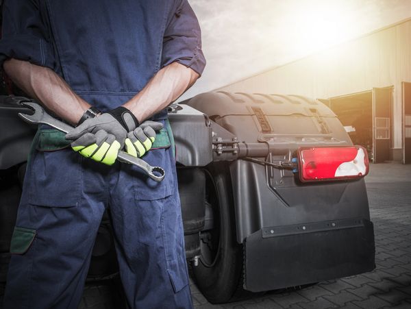 Mechanic in gloves holding a wrench behind his back near a truck.