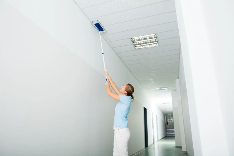 Young Woman Cleaning The Ceiling With Mop On The Corridor Of The Building
