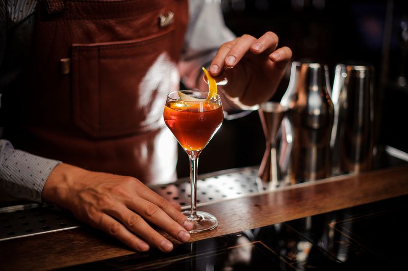alcohol drinks, people and luxury concept - bartender with glass and lemon peel preparing cocktail at bar