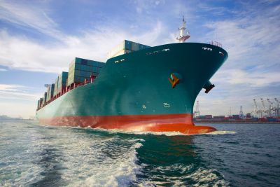 Large cargo ship carrying containers sails near a port under a blue sky.