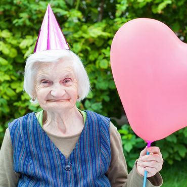Elderly woman wearing a party hat holding a pink heart-shaped balloon outdoors.