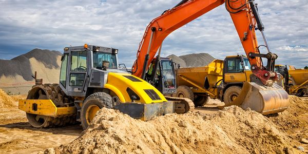 Orange excavator and yellow dump truck at a construction site with gravel piles.
