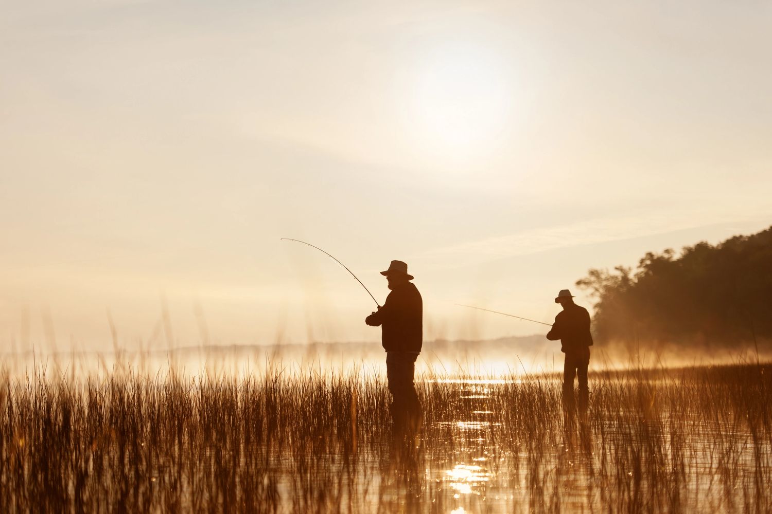 Two people fishing in a misty lake during sunrise.