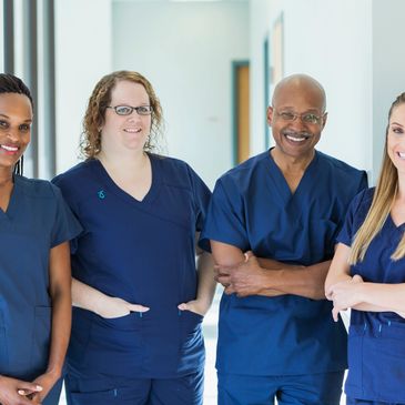 Four healthcare professionals in blue scrubs smiling in a hallway.