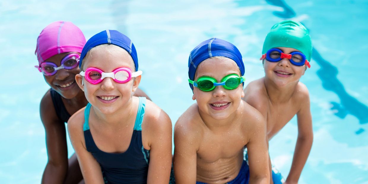 Four children in swim caps and goggles smiling at the poolside.