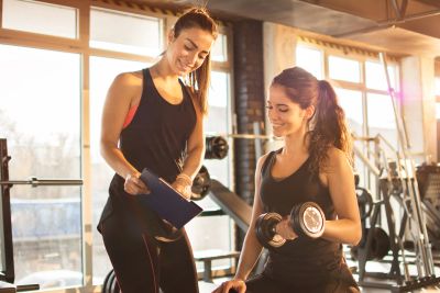 Two women in workout attire exercise with dumbbells in a gym.