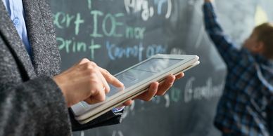 Teacher uses tablet while student writes on chalkboard in classroom.