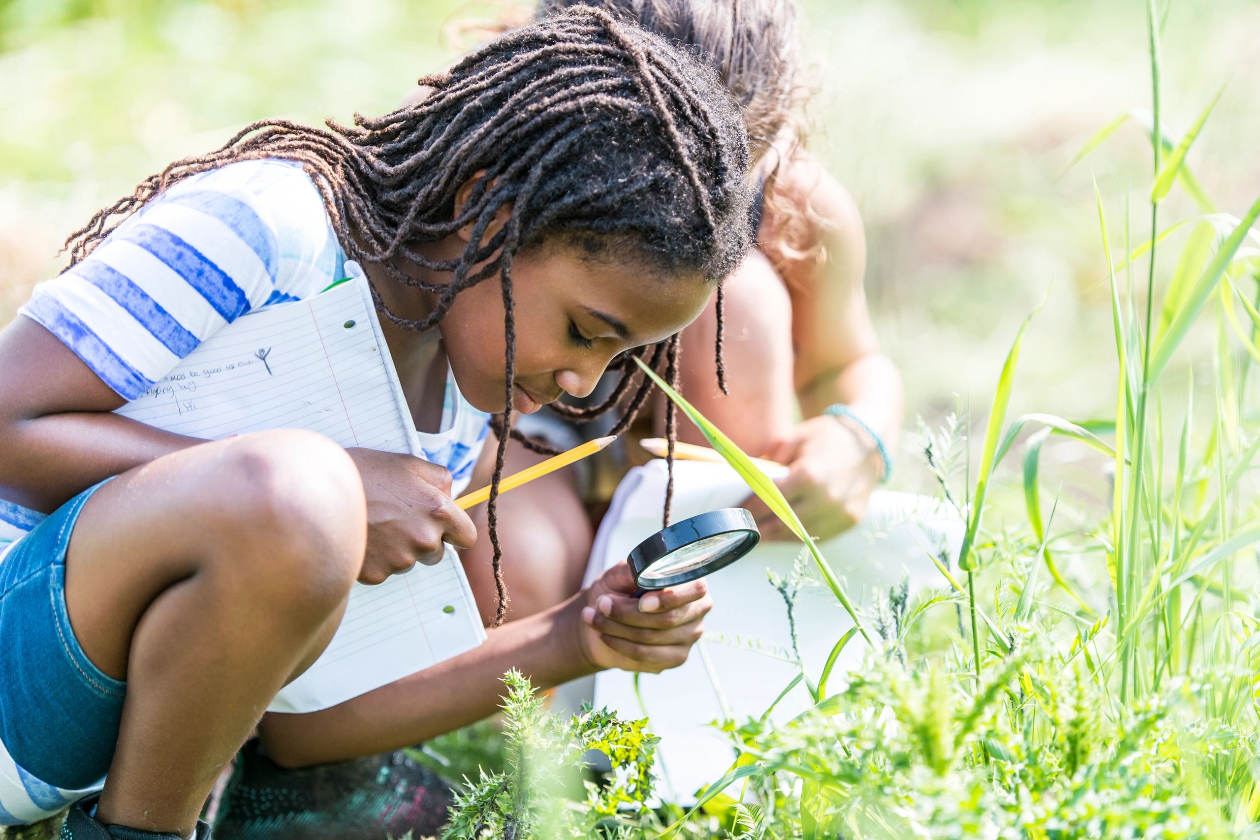 Happy young girl outside, holding a magnifying glass, crouching down to observe grasses and thistles