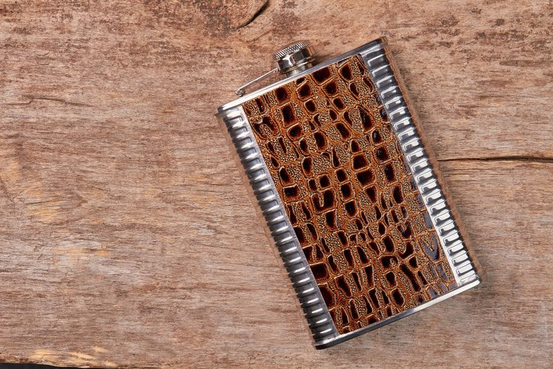 Stylish brown hip flask, wooden background. Steel jhip flask as souvenir.