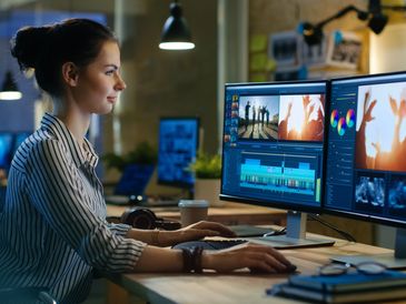 A woman editing video on dual monitors in a modern office.