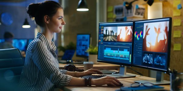 A woman editing video on dual monitors in a cozy workspace.