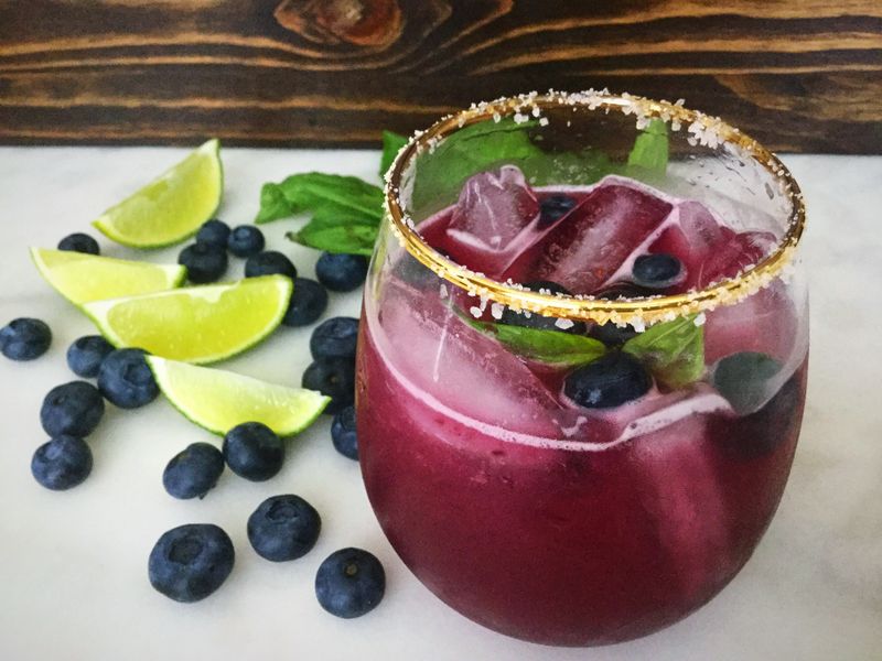 Margarita cocktail and ice in a salt-rimmed shown with blueberries, lime wedges and basil leaves