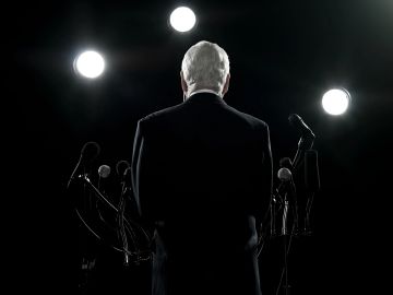 Elderly man standing behind microphones under bright lights, facing away.