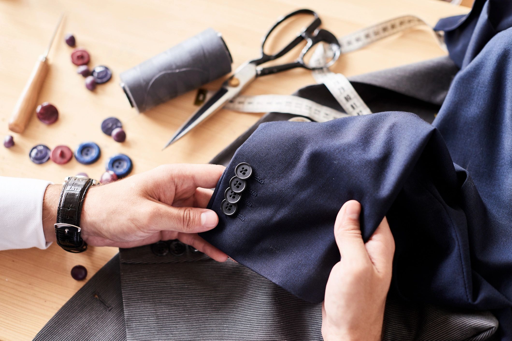 Tailor inspecting sleeve buttons on a navy blue suit jacket with sewing tools nearby.