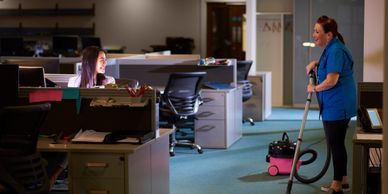 Nighttime office scene with a janitor vacuuming and an employee smiling at her.